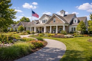 Exterior photograph of an upscale 55+ active adult community in Lancaster County Pennsylvania, showing a clubhouse building with colonial architecture, stone and siding exterior, covered porch with rocking chairs, American flag, beautifully landscaped grounds with flowering gardens, walking paths visible, golf cart path in foreground, blue sky with white clouds, warm and inviting atmosphere. Professional community marketing photography style. No people visible but suggesting active lifestyle.